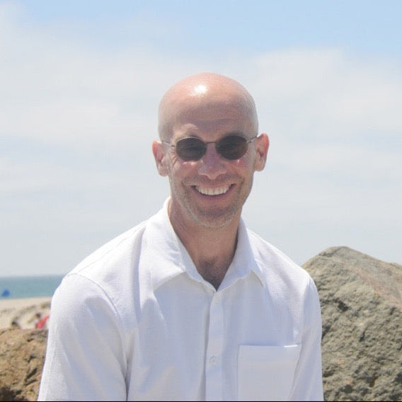 Man wearing sunglasses and a white shirt on a rocky beach with a clear sky.